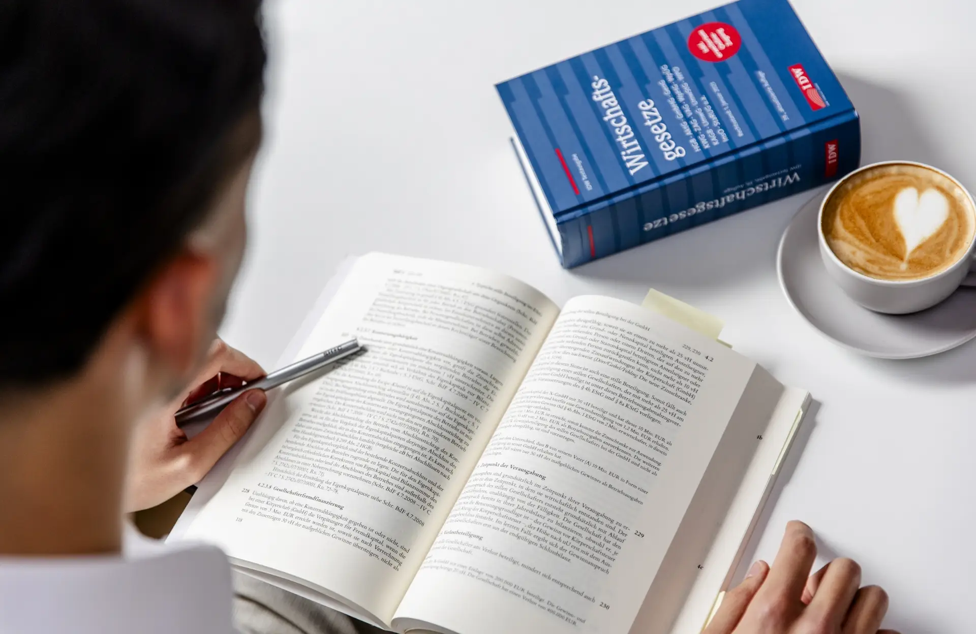 Steuerberater liest in einem Buch für Wirtschaftsgesetze; eine Tasse Cappuccino mit Milchschaumherz gibt der Szene eine sympathische Note.
