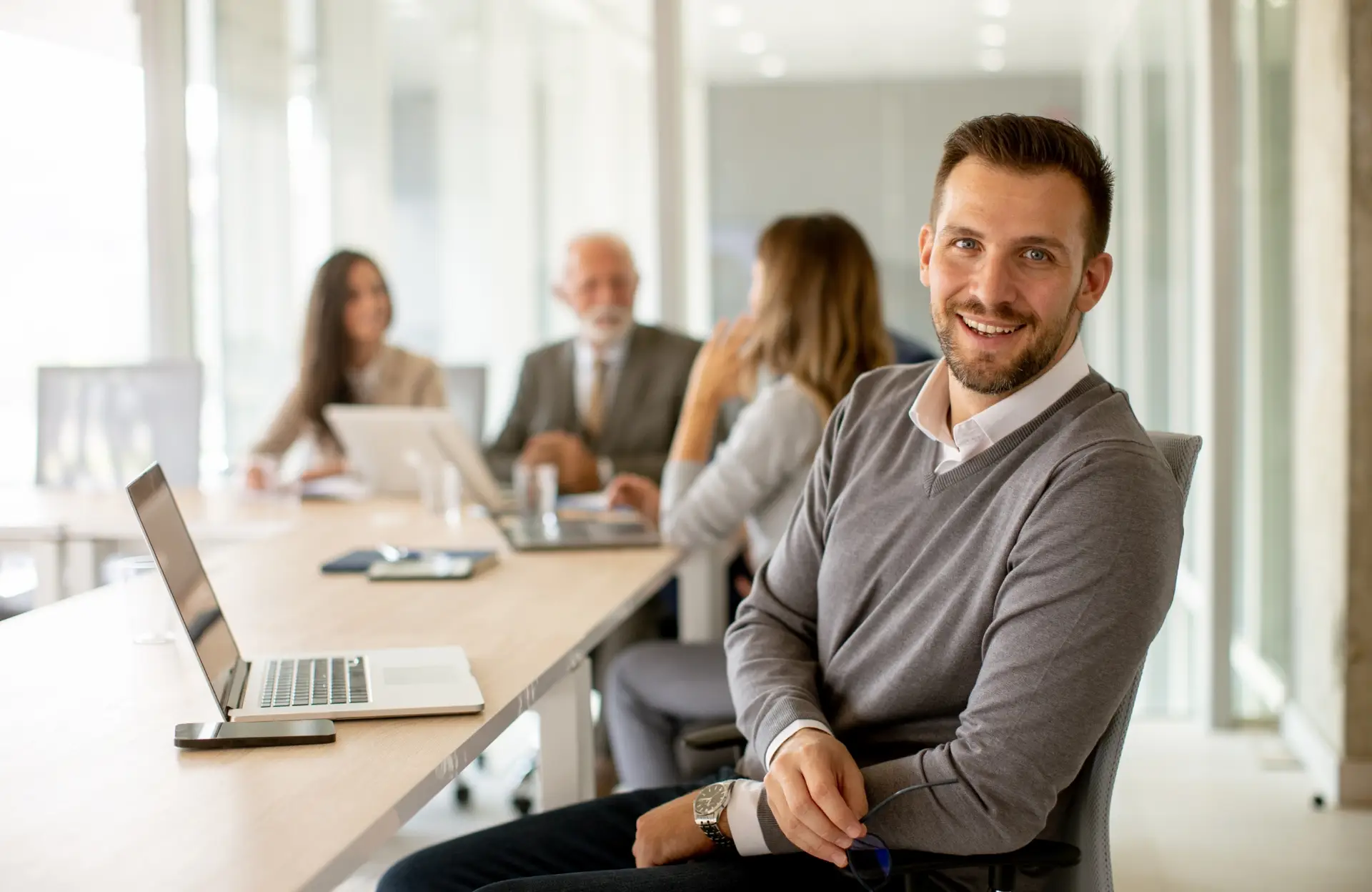 Sympathisch lächelnder Berater am Konferenztisch mit zwei jungen Damen und einem betagten Herrn.