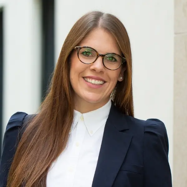 Bachelor of Arts (B.A.) Bianca Ott in blauem Blazer und heller Bluse vor heller Hauswand.