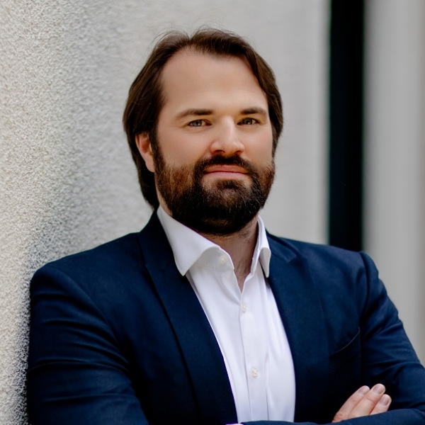 Steuerfachangestellter Maximilian Wittmaier in blauem Jackett und weißem Hemd vor heller Hauswand.