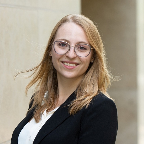 Sara Sacher mit langen, blonden Haaren und blauem Blazer sowie weißer Bluse vor Hauswand